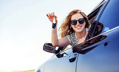 A woman holding a car key with her head and arm sticking out of the driver's side window of a car.