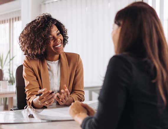 A pair of women sitting across from each other at a table, smiling while appearing to discuss the contents of a binder that is sitting on the table