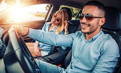 A picture looking in through a car's front driver side window at a man smiling and driving with a woman smiling in the passenger side front seat.