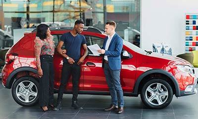 Three people standing in front of a red SUV, appearing to be having a discussion about the contents of some papers one of them is holding.