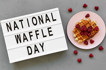 A small marquee with text reading national waffle day laid on it's back, sitting by a plated waffle that is topped with and raspberries.