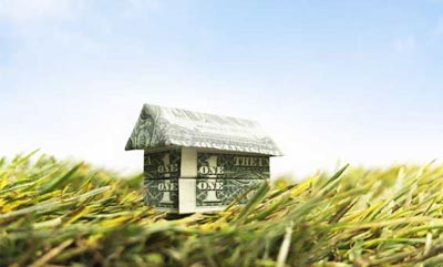 A dollar bill has been folded into the shape of a home & it's sitting on green grass with a blue sky backdrop