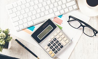 A calculator sits on a desk alongside a pen, keyboard and black framed glasses