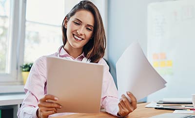 A smiling woman, looking at a page from stacks of papers she is holding in her hands.