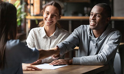 A smiling couple sitting behind a desk, and one of them is shaking hands with someone on the other side of the desk