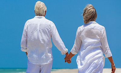 An older couple is holding hands as they walk away on a sandy beach.