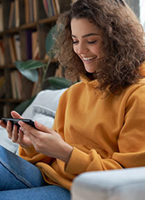 A woman is sitting on a sofa, holding a cellphone that she is looking at an smiling.