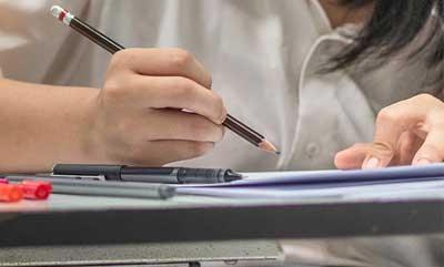 A close up picture of a person's hand holding a pencil over a paper that is on a desk