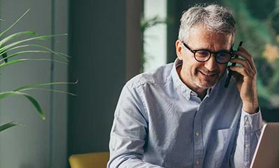 A man is sitting a a desk, smiling, looking at a laptop and listening to a phone call on his mobile device