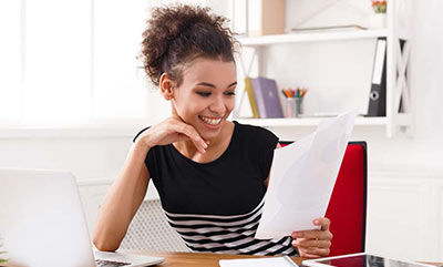 A smiling young woman sitting at a desk, holding a piece of paper that she is looking at.