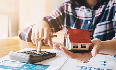 A person pushing a button on a calculator and holding a small model of a house in the other hand.