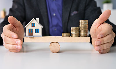 A scale with a house on the left side and a few stacks of coins on the right side.