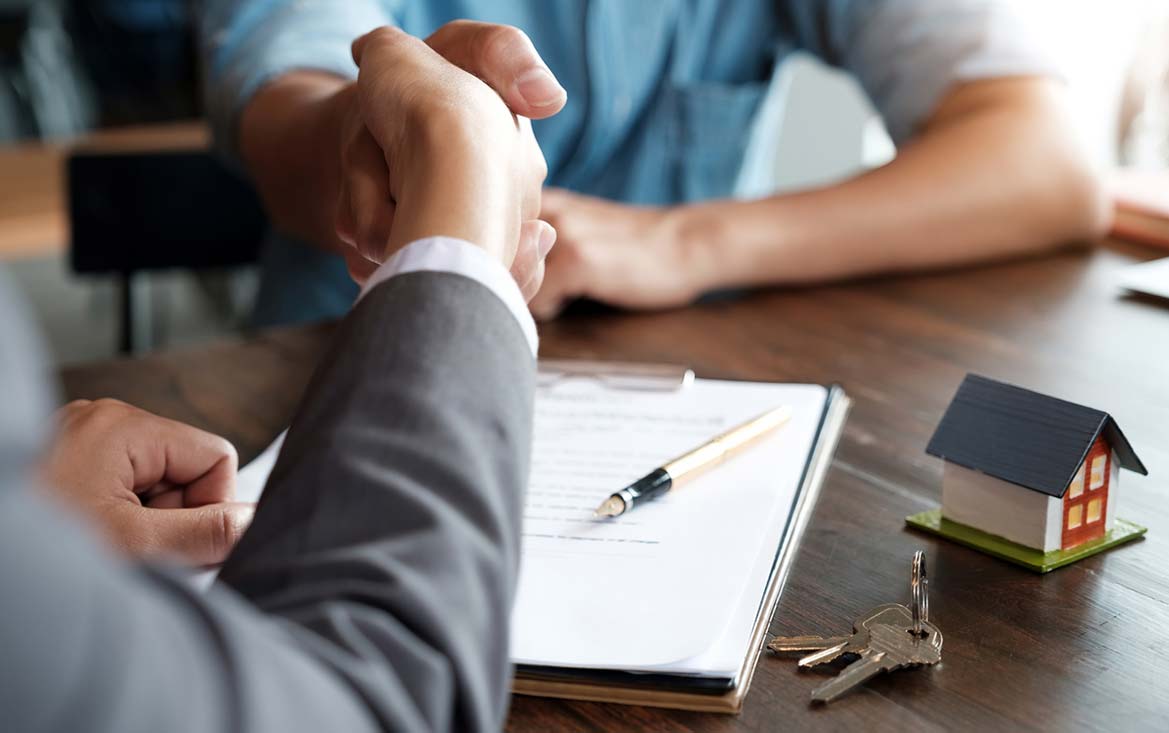 A close of the handshake of two individuals who are sitting across a desk from each other with a few table top items in view, including a clipboard with papers and a pen on it, a small model of a home, and a set of keys